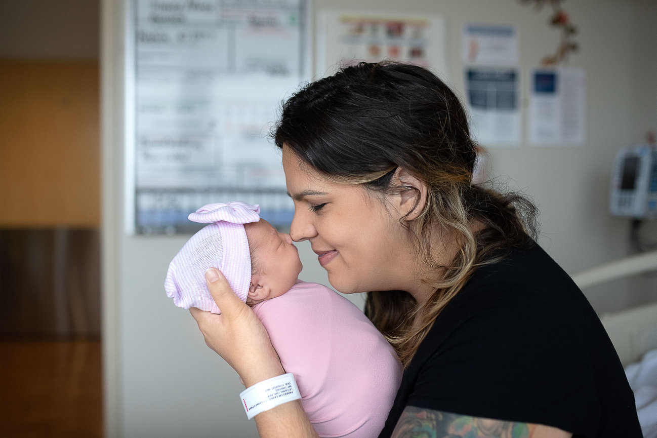 In a cozy hospital room in Huntley, Illinois, a woman with a hospital wristband gently touches noses with her newborn, who is snugly wrapped in a pink blanket and hat. Both are smiling warmly, capturing an intimate moment of joy and connection.