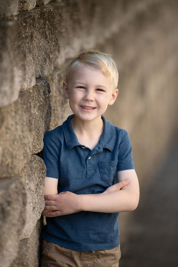 Smiling boy with blonde hair, wearing a blue polo shirt and khaki pants, stands with arms crossed against a stone wall background, perfectly capturing the charm of timeless family photos.