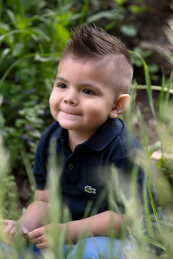 A young child with short, spiky hair sits outdoors among lush greenery. Wearing a dark blue polo shirt, the child gazes thoughtfully to the side—an ideal moment captured for family photography near me.