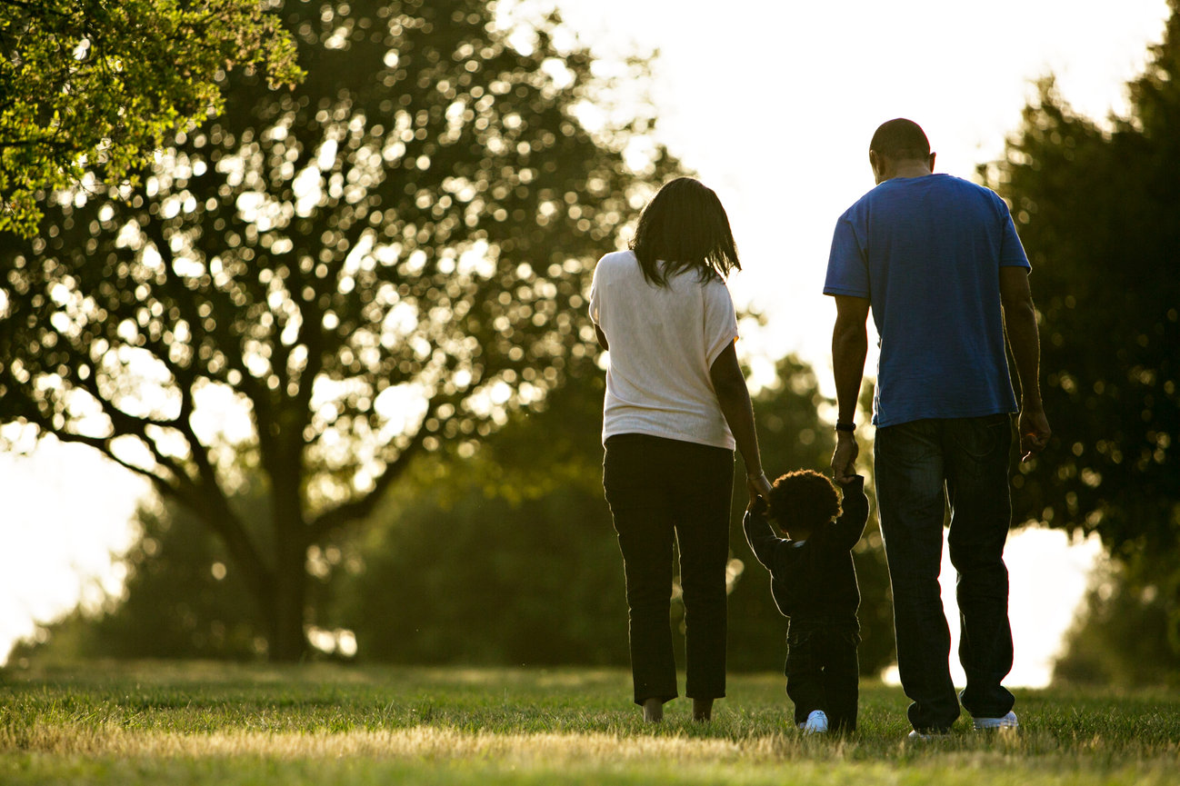 African American, Black parents walking away with their toddler, holding his hands, silhouetted with trees and pastoral background