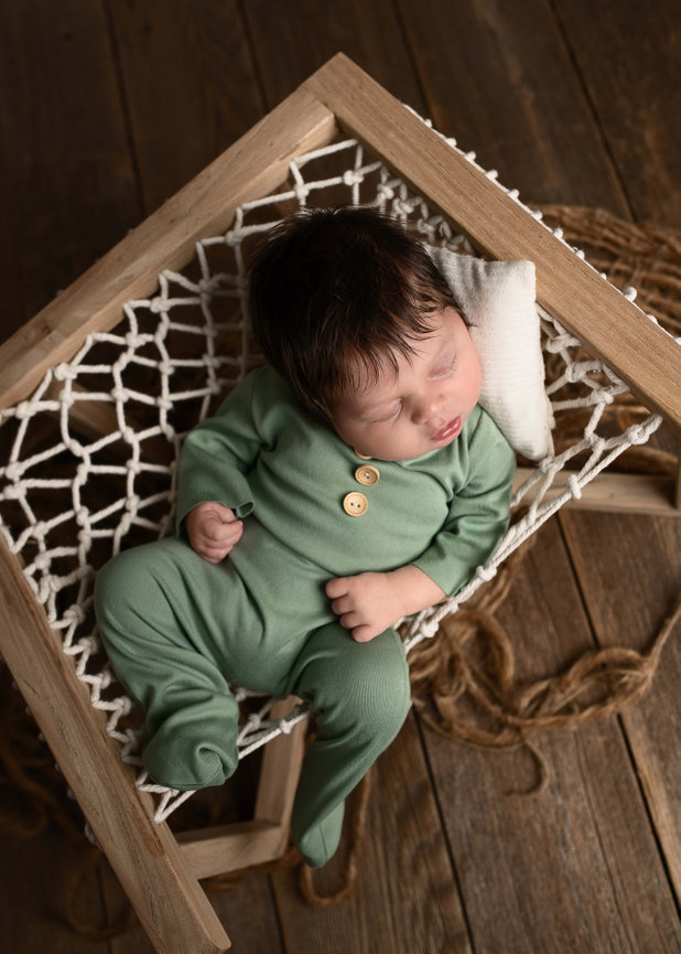 A baby in a green onesie sleeps peacefully on a wooden hammock with a small white pillow. The hammock is netted, and the baby has dark hair. The background is wooden, adding a rustic feel to the scene.