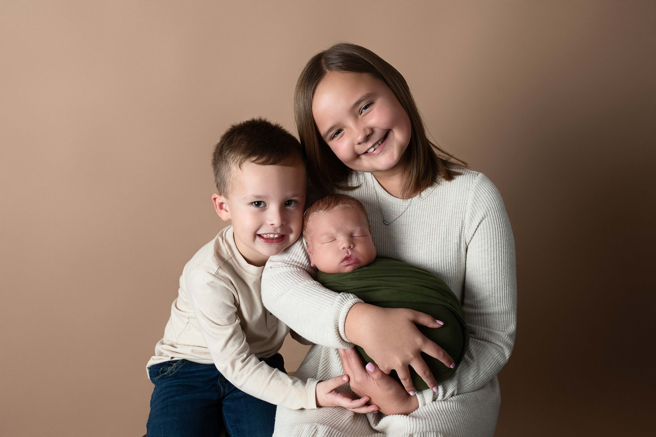 A young girl and boy sit closely together, smiling warmly at the camera. The girl gently cradles a sleeping newborn wrapped in a green blanket. This charming sibling foto is set against a soft, neutral background.