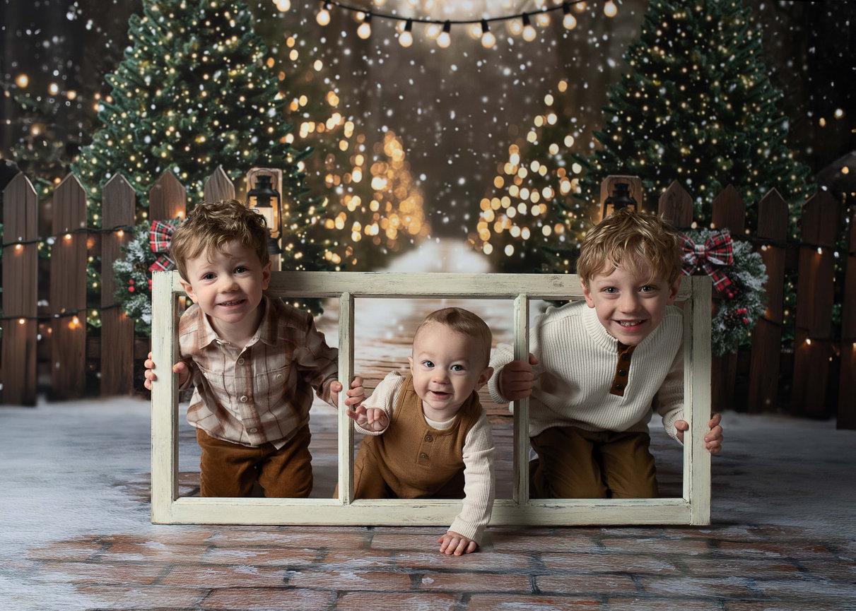 Three young children, including their nine-month-old sibling, joyfully lean through a wooden frame against a festive, snowy backdrop. String lights and decorated Christmas trees enhance the holiday atmosphere. They wear cozy outfits and smile happily