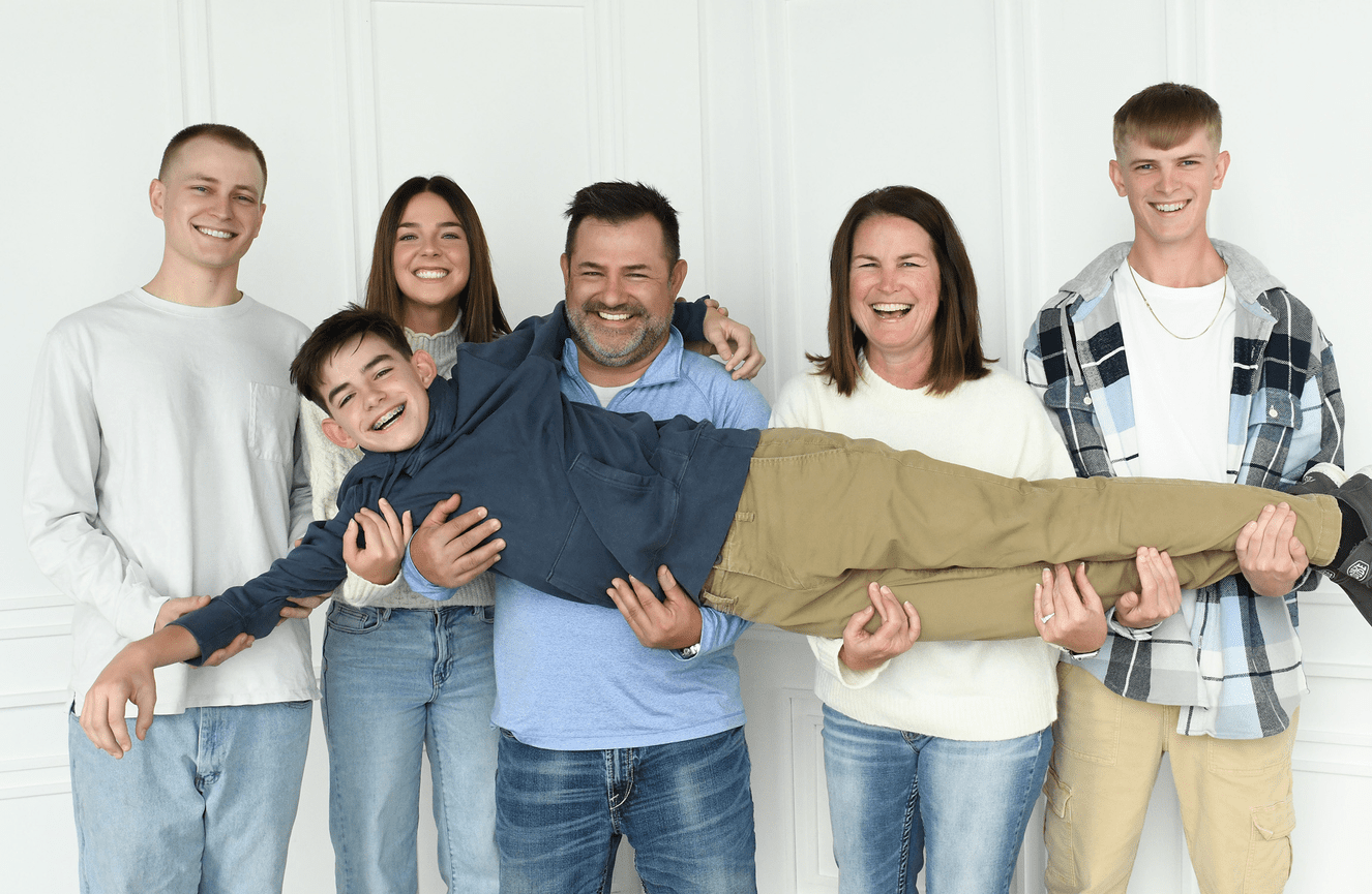 A fun family photo where everyone is standing in a row while the youngest boy has been picked up and held across horizontally while they all laugh.