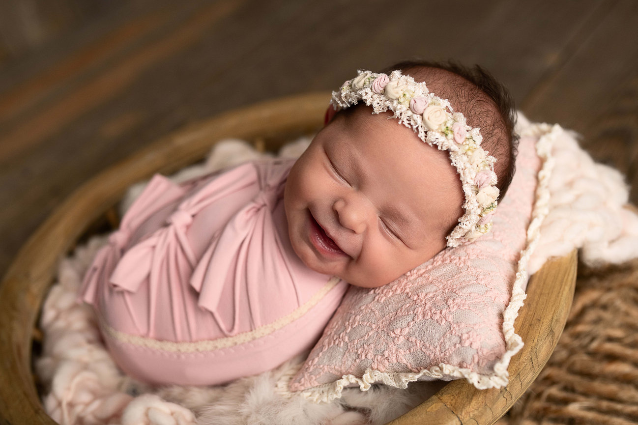 A newborn baby is swaddled in a pink wrap, resting on a textured pillow inside a wooden bowl in McHenry, Illinois. The baby, adorned with a floral headband and smiling with closed eyes, is surrounded by a soft blanket that falls on the wooden floor.
