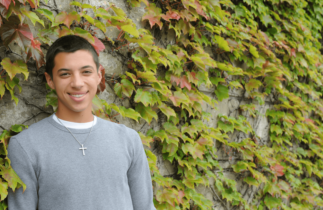 A guy wearing a cross necklace standing against a wall of autumn ivy.