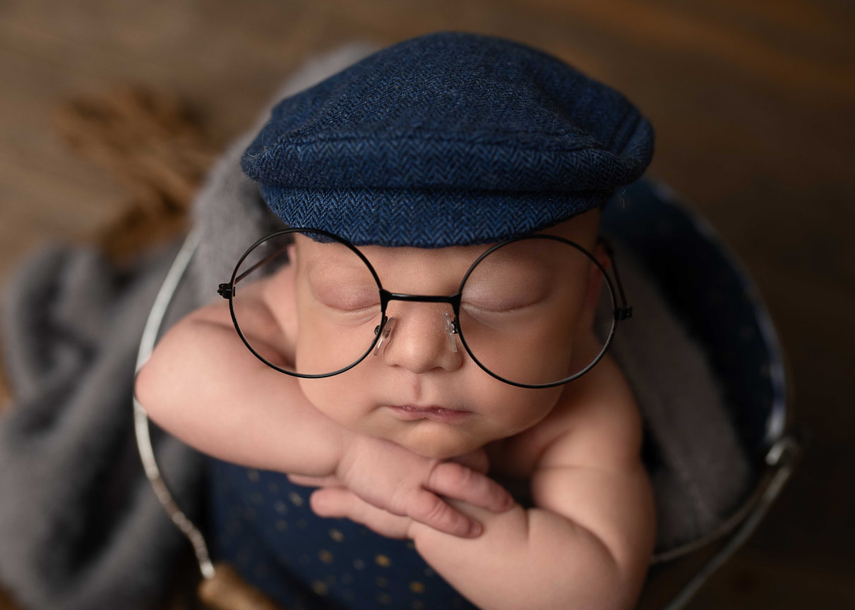 Adorable newborn baby wearing oversized round glasses and a blue flat cap, peacefully sleeping with arms crossed in a cozy bucket setup – vintage-style newborn photography in Barrington, Illinois