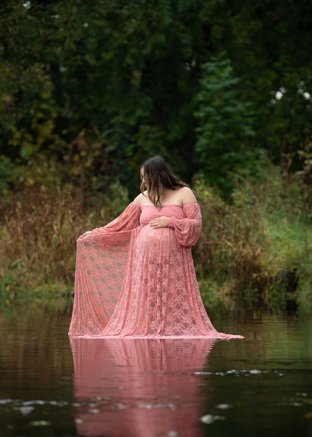 Pregnant woman in pink lace gown stands in shallow water surrounded by lush greenery.