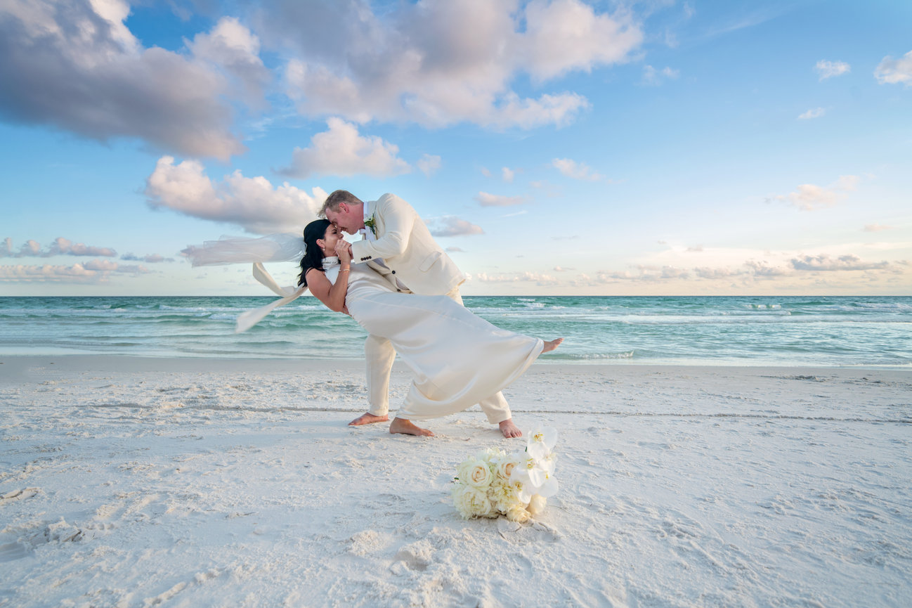 Couple in wedding attire dancing on a sandy beach with a bouquet, ocean waves, and a cloudy blue sky.