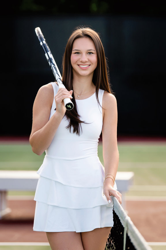 Westosha Central senior holding a tennis racket, standing by a net on a tennis court, wearing a white dress for her senior pictures.