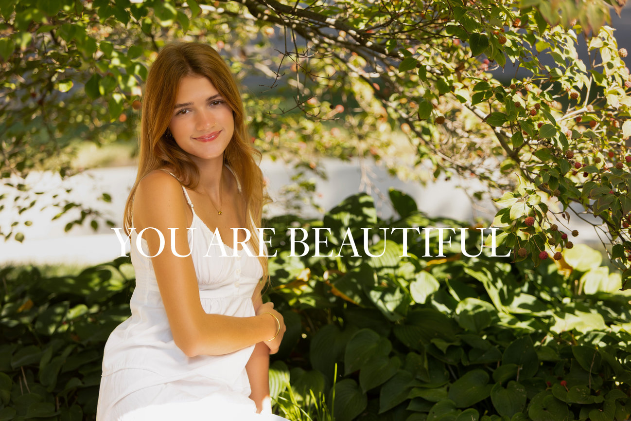 Woman in white dress sitting outdoors amidst greenery with text You Are Beautiful overlaid.