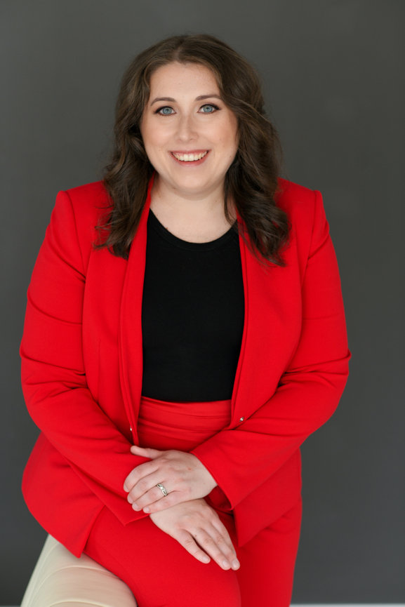Woman being photographed for updated headshots in Dublin Ohio, in a red suit sitting against a gray background, smiling confidently.
