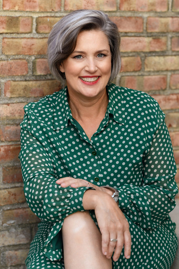 Woman with short, silver hair smiles while seated against a brick wall, wearing a green polka dot blouse for professional headshots in Worthington Ohio