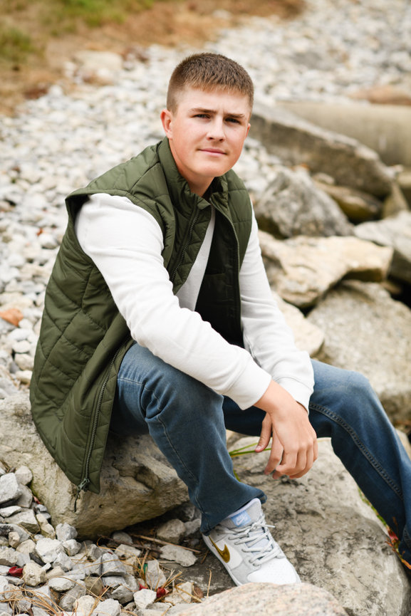 Teenage boy in a white long sleeved shirt with a green vest sitting on large rocks for senior portraits in Dublin OH.