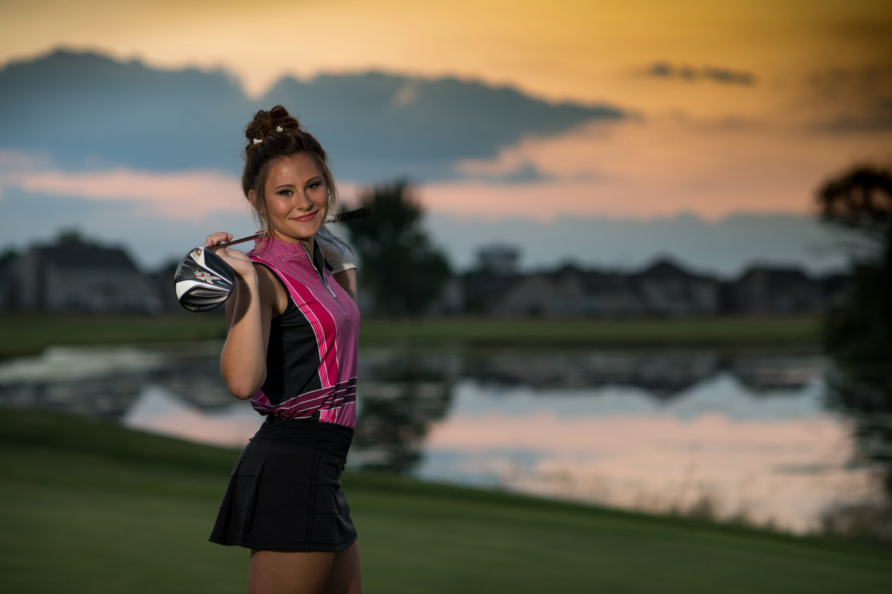 Golfer in a stylish outfit poses with a driver club at sunset on a golf course.
