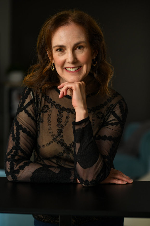 Smiling woman in a sheer black top resting chin on hand, seated in a stylish indoor setting for updated headshots and dating profile photos in dublin ohio.