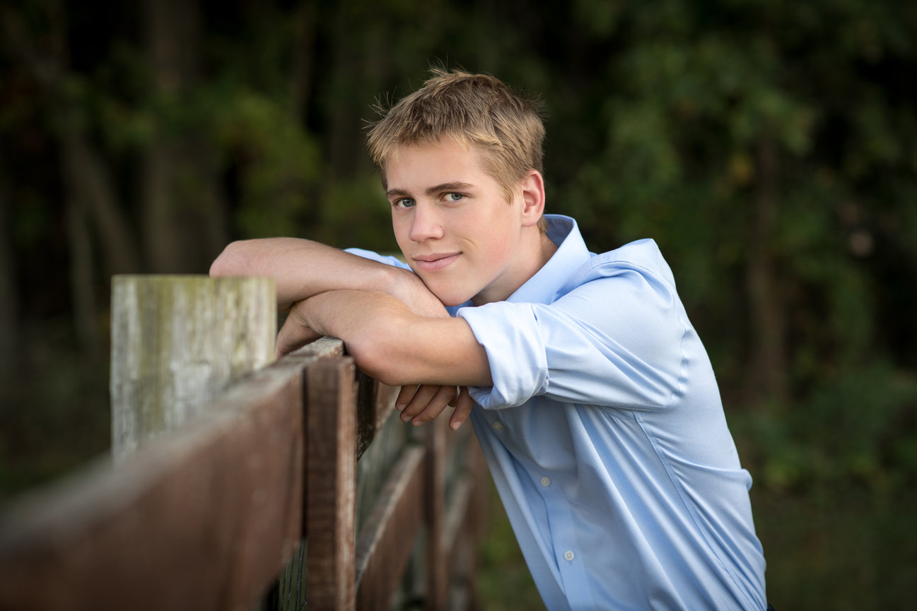 Teen boy in blue shirt leaning on a wooden fence, smiling, with wooded area in the back for senior pictures in Dublin Ohio.