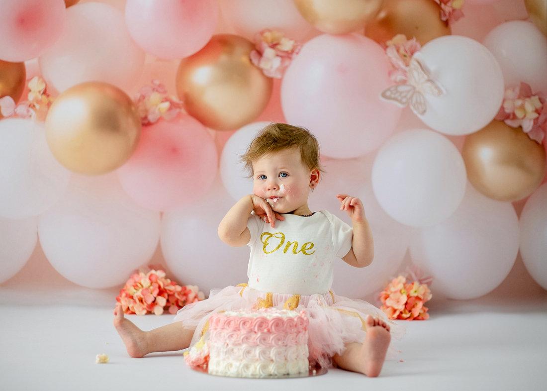 Baby girl in a white “one” shirt and tutu enjoying her cake during a pink and gold balloon-wall cake smash by Cake Smash by Chris in Point Cook.