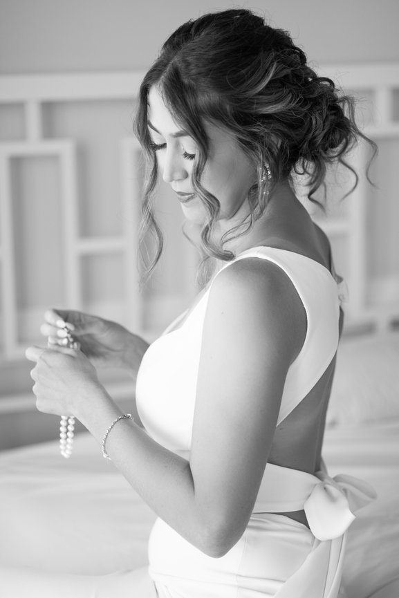 Bride in white dress holding pearl necklace, hair styled in curls, standing indoors.