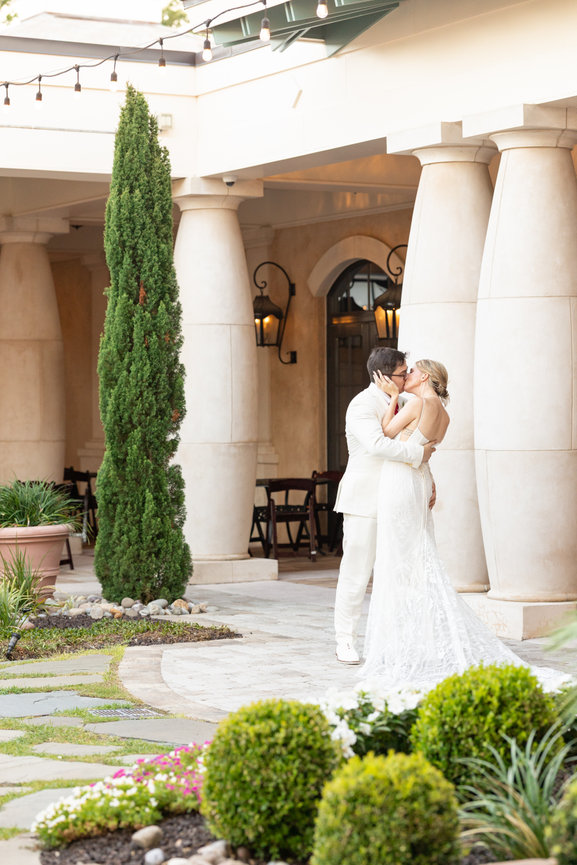 Bride and groom kissing outdoors near elegant columns and greenery.